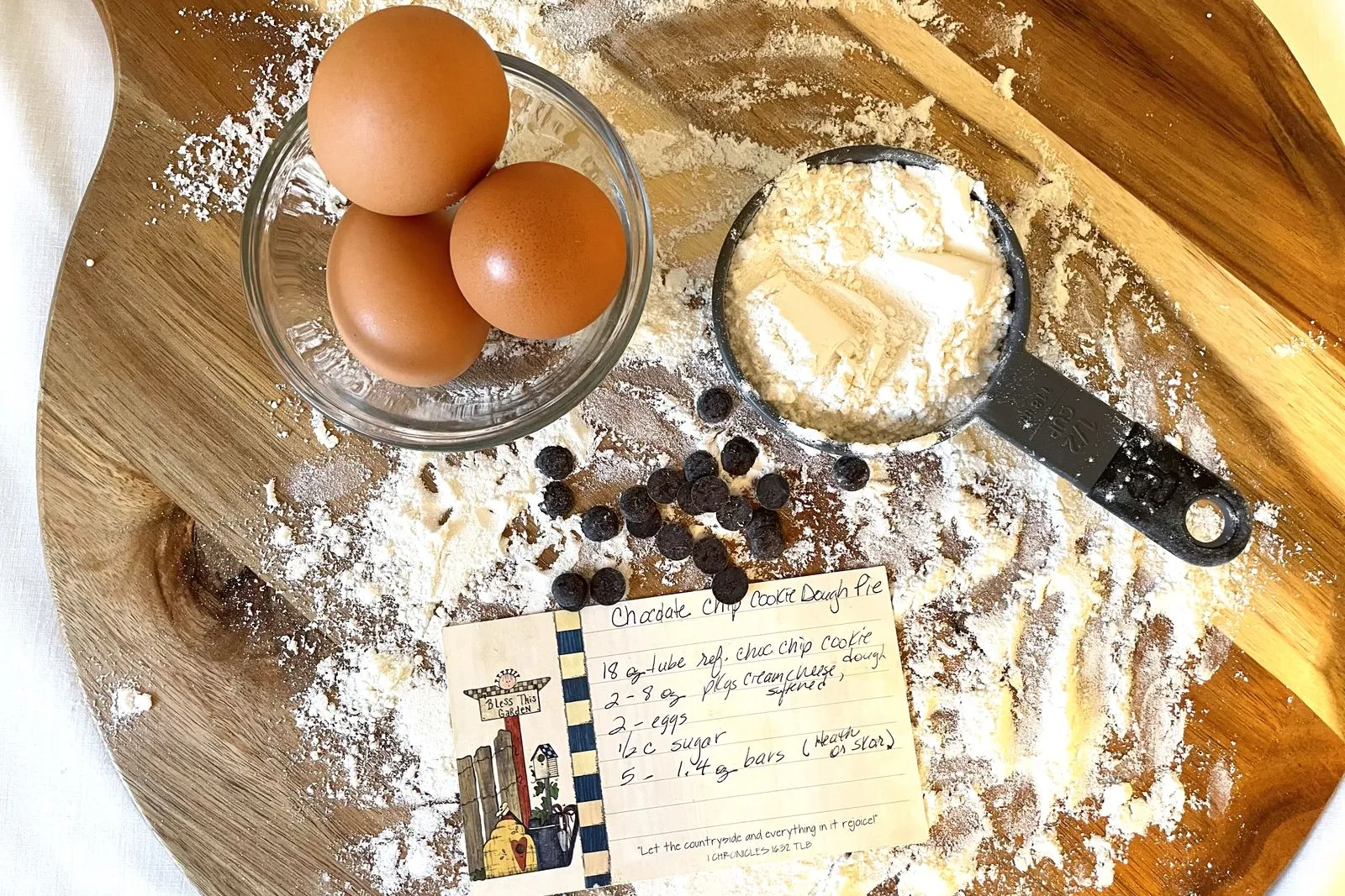 Three brown eggs in a glass bowl, a measuring cup of flour and a handwritten recipe card on a floured wooden board.