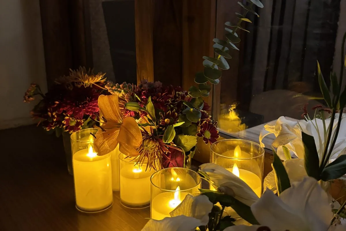 Lit candles and a vase of lilies and autumn flowers on a table beside a window in the evening.