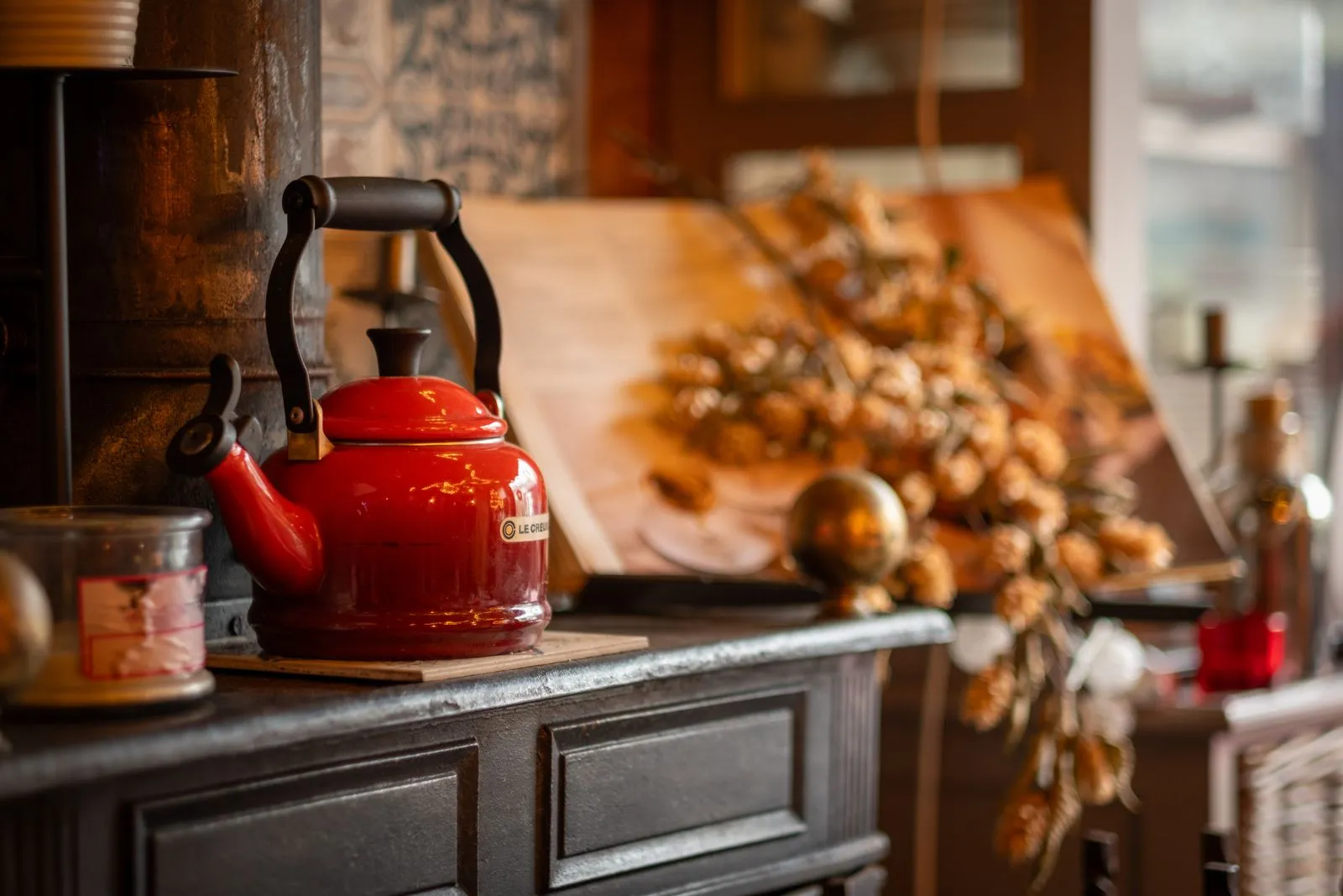 A red enamel kettle on a dark wooden cupboard in a kitchen, with dried autumn decorations behind it.