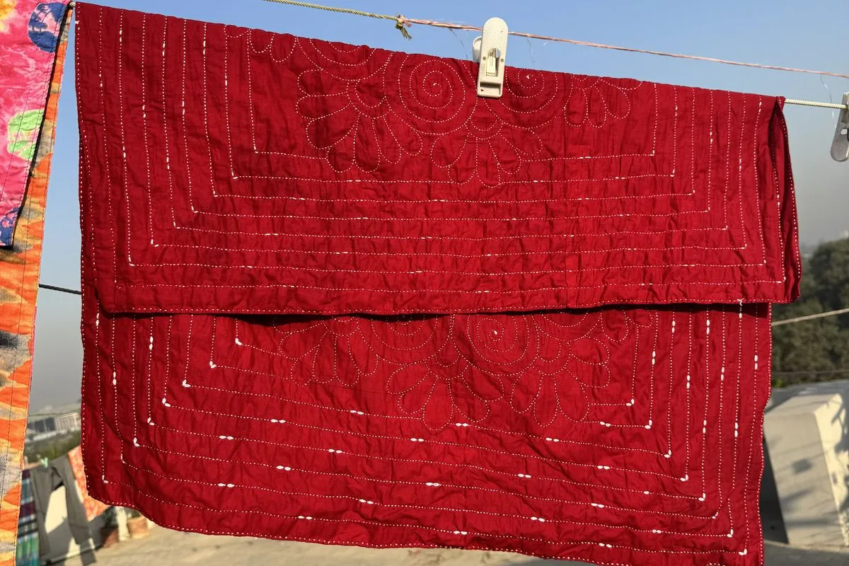 A deep red hand-stitched quilt pinned to a clothesline on a sunny rooftop, its shadow falling on the tiles.