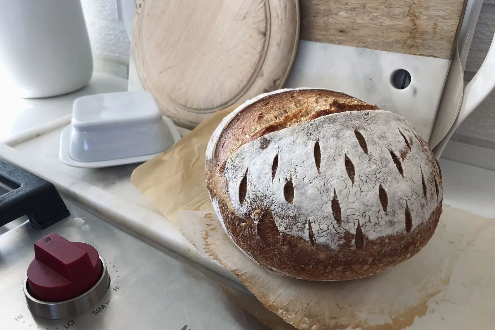 A scored round sourdough loaf cooling on a marble kitchen counter beside a bread board and a butter dish.